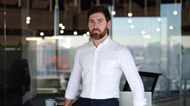 Modern bir ofisteki kendine güvenen işadamı gülümser ve onaylar. Erkek mutludur ve işten hoşlanır. Doğal ışık, cam duvarlar. Başarı portresi iş adamı