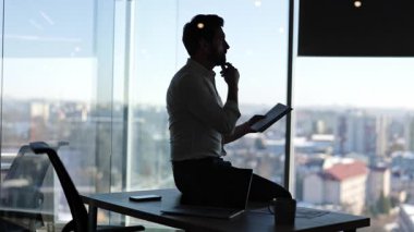 Düşünceli iş adamı, modern ofisteki masasında oturan defterine yazıyor. Erkek girişimci girişim planlama projesi. Büyük şehir ofisindeki iş adamı silueti..
