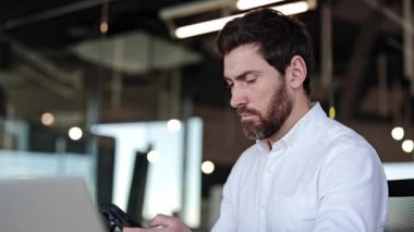 Modern ofiste telefonla konuşan odaklanmış bir işadamı. Beyaz tişörtlü sakallı adam akıllı telefonla çalışır, bilgisayarı da arka planda..