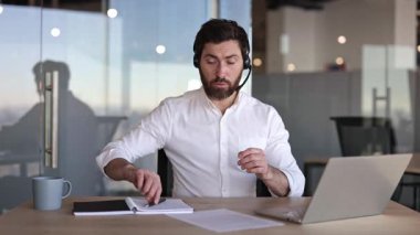 Kulaklık takan kendine güvenen iş adamının modern ofiste dizüstü bilgisayarında görüntüsü var. Genç erkek profesyonel konuşuyor, müşteriyle bir şey tartışıyor ya da adayla röportaj yapıyor..
