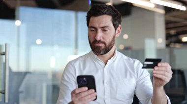 Sakallı başarılı bir iş adamı akıllı telefondan online ödeme yapıyor. Modern bir ofiste kredi kartıyla ödeme yapan mutlu bir işadamı..