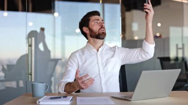 Ofiste daha iyi bir cep telefonu şebekesi kurmaya çalışan hayal kırıklığına uğramış iş adamı. Masada oturan sakallı erkek akıllı telefona bakıyor ve kötü bir bağlantıyla kolunu kaldırıyor..