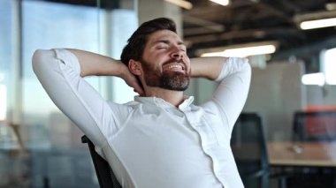 Rahat iş adamı elleri başının arkasında, elleri kapalı, gülümseyerek ofis sandalyesine yaslanıyor. Erkek şirket çalışanı, yönetici yönetici derin bir nefes alıyor ve sakinlikten hoşlanıyor..