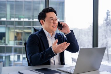 Başarılı iş adamı telefonda konuşuyor ve iş görüşmesi sırasında el kol hareketi yapıyor. Profesyonel gibi giyinmiş, dizüstü bilgisayarla masaya oturmuş. Kurumsal ofis geçmişi, başarı ve iletişim.