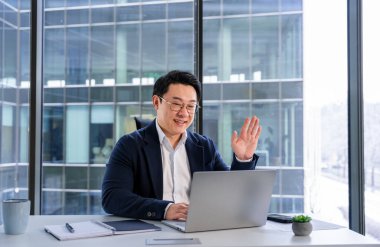 Modern ofisteki profesyonel işadamı bir video çağrısına katılıyor, pozitifliğini ve bağlantısını dile getiriyor. Üretkenlik ve profesyonel etkileşim, işyeri verimliliğinin artırılması.