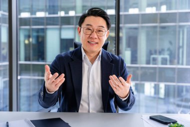 Kendine güvenen Asyalı işadamı şık günlük kıyafetler giyer, açıklama yapar, büyük cam pencereli modern bir ofiste oturur, profesyonellik ve etkili iletişim becerileri yayar..