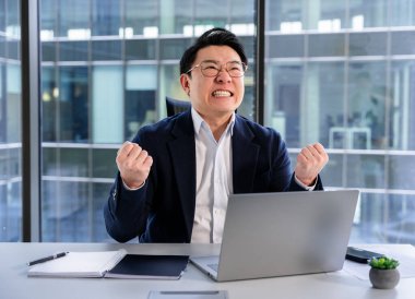 Modern bir ofiste profesyonel bir Asyalı işadamı, gözlük takıyor ve resmi kıyafetler giyiyor, dizüstü bilgisayar kullanırken stres yapıyor. Arka plan cam pencerelerden karlı bir kentsel ayar gösteriyor.