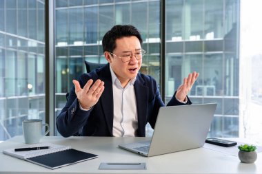 Profesyonel Asyalı işadamı, modern bir ofis masasında çalışırken hayal kırıklığını dile getiriyor, iş gereksinimleriyle çevrili. Kentsel yapı arka planı çalışma alanı ayarlarını tamamlar.