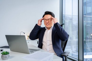 Ofis ortamında başı tutan profesyonel bir işadamı, stres ya da hayal kırıklığını ifade ediyor. Bilgisayarlı ve pencerelerden görünen şehir manzaralı modern çalışma alanı kurumsal ortamı vurguluyor.