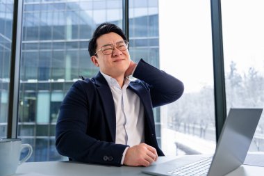 Ofis ortamında profesyonel bir Asyalı işadamı, görevler sırasında fiziksel rahatsızlığını gösteriyor, bir masa başında dizüstü bilgisayar ve modern mimarinin pencere manzaralı iş kıyafetleri giyiyor..