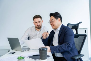 Profesyonel işadamları modern bir ofiste iş birliği yaparak, dizüstü bilgisayarlar ve diğer iş gereklilikleriyle dolu bir masada otururken belgeleri tartışır, takım çalışması, odaklanma ve kurumsal bir ortam sunar..