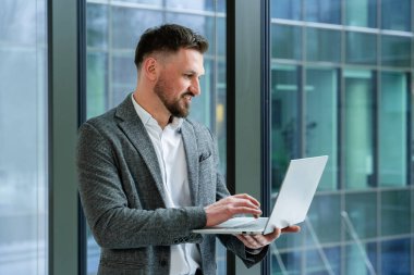 Büyük bir ofis penceresinin yanında dizüstü bilgisayar kullanan başarılı bir iş adamı, odak ve profesyonelliği ifade ediyor. İş ortamı, doğal ışık verimliliğini artıran doğal ışık ortamıyla çağdaş.