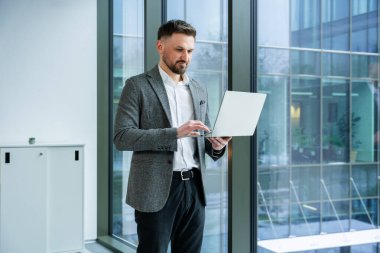 Modern ofis ortamında büyük pencerelerin yanında dizüstü bilgisayar kullanan profesyonel bir işadamı şık bir takım elbise giyer ve çağdaş bir iş yerinde odak ve üretkenliğini ifade eder..