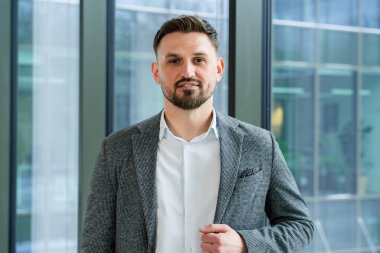 Modern ofis ortamında profesyonel bir işadamı, takım elbise ve gömlek giyiyor, kendine güvenini ve duruşunu büyük bir pencerenin yanında gösteriyor, kentsel çevre, kurumsal tutum.