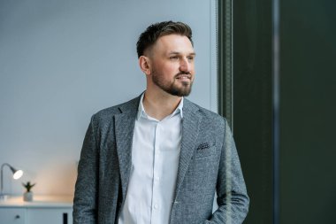 Modern bir ofiste pencerenin önünde duran profesyonel işadamı şık kıyafetler giyiyor, kendine güven ve başarı yayıyor. Parlak doğal aydınlatma ve çağdaş alan iş atmosferini vurguluyor.