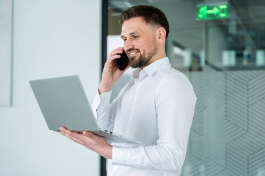 Zarif beyaz gömlekli kendine güvenen iş adamı modern bir ofiste dizüstü bilgisayar ve telefonla birden fazla iş yapıyor, verimlilik, iletişim ve işyeri profesyonelliğini neşeli bir tavırla sergiliyor..