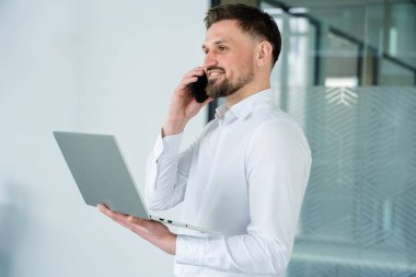 Profesyonel iş adamı resmi giyiniyor. Laptop ve akıllı telefon ile modern bir ofis içinde verimliliği ve başarıyı temsil ediyor..