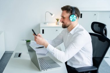 Genç bir adam modern bir masada dizüstü bilgisayarla cep telefonu kullanıyor ve şık kulaklıklar takıyor. Modern ortamlarda iletişim, teknoloji ve ofis verimliliğini temsil eder.