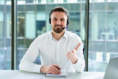Profesyonel işadamı kulaklık takıyor, modern bir ofis masasında çevrimiçi bir toplantıya katılıyor, odaklanmış ve üretken bir çalışma gününde güven ve iletişim becerilerini canlandırıyor..