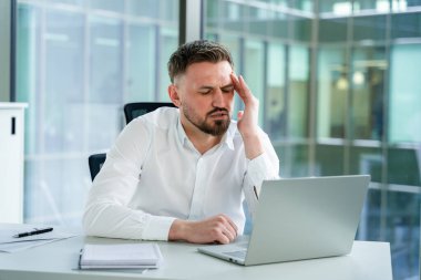 Modern ofis ortamında dizüstü bilgisayar kullanırken hayal kırıklığına uğramış görünen profesyonel iş adamı. Beyaz bir gömlek giyiyor ve ofis malzemeleriyle çevrili..