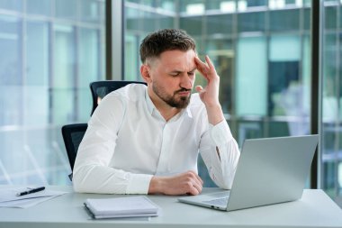 Modern bir ofiste beyaz gömlek giyen profesyonel bir adam dizüstü bilgisayarla çalışırken stres yaşıyor. Sahne, kurumsal bir ortamda iş sorunları üzerine hayal kırıklığı ya da endişeyi yakalıyor.