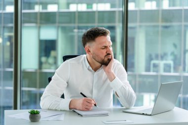 Modern bir ofiste oturan profesyonel bir adam, resmi kıyafetler giyiyor, deftere yazıyor, odaklanmış ve acı dolu ifadeler sergiliyor. Pencereleri ve beyaz bir masada dizüstü bilgisayarı olan parlak çalışma alanı.