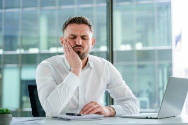 Modern bir ofis masasında acı çeken profesyonel bir işadamı, rahatsızlık ve hüsranı yansıtıyor. Resmi gömlek giyimi profesyonel çevrenin güvenilirliğini artırır.