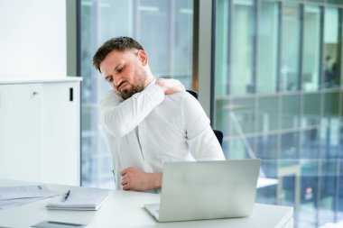 Erkek, kurumsal bir ofiste rahatsızlık yaşıyor, omuz ağrısı çekiyor, dizüstü bilgisayar ve kağıtlarla otururken resmi kıyafetler giyiyor, işyerindeki sağlık sorunlarını ve stres atılımını vurguluyor..