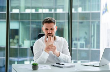 Ofisteki profesyonel bir adam, bileğini rahatsızlıkla tutuyor. Dizüstü bilgisayarla ve belgelerle modern çalışma yerinde oturuyor. Cam bir pencerenin yanında, işyeri sağlık sorunlarını resmediyor..