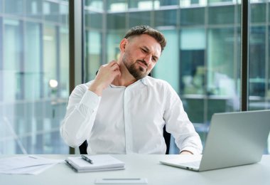 Modern bir ofiste profesyonel bir adam dizüstü bilgisayar kullanırken boyun rahatsızlığı çekiyor. İş kıyafetleri, iş yeri sorunları ve kurumsal bir ortamda ergonomik sorunlar..