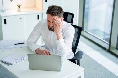 Profesyonel işadamı modern bir ofiste oturuyor ve dizüstü bilgisayarla çalışırken stresli görünüyor. Evraklar ve defterler onu çevrelemiş, stresli ve yoğun bir çalışma ortamını resmediyor. İş konsepti.