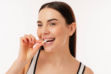 Gülümseyen kız, temiz, minimalist bir ortamda ağzının yanında beyaz bir hap tutuyor. Sağlık, sağlık ve beslenmeyi temsil eder..