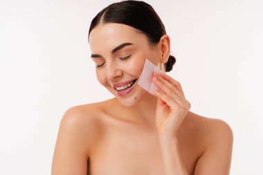 Taze ve ışıl ışıl cilt görünümünü, cilt bakımını ve sağlıklı yaşam tarzı kavramlarını korumak için lekeleme kağıdı kullanan bir kız. Tarafsız arka plan, konu etkinliği ve temizliğine odaklanmayı artırır.