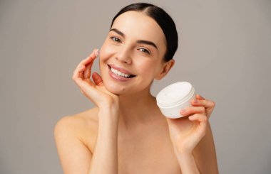 Gülümseyen kadın elinde beyaz bir teneke kutu dolusu cilt bakımı, güzellik ve kişisel bakım malzemesi ile temiz ve tarafsız bir arkaplan tutuyor. Cilt bakımının parlak ve neşeli bir temsili.