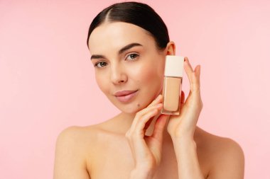 Zarif bir birey, pastel pembe bir arka plana karşı bir şişe deri temeli sunuyor. Güzellik ve cilt kavramlarını vurguluyor..