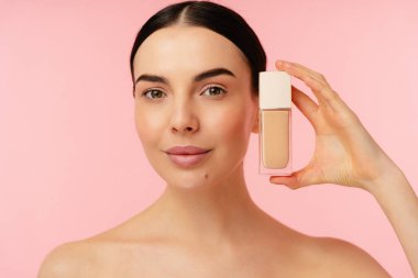 Canlı pembe bir arkaplana karşı kozmetik bir vakıf kabı sergileyen bir şahsın portresi.