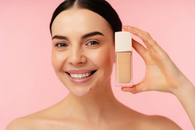 Elinde sıvı bir fondöten şişesi olan gülümseyen bir kız, kendine güvenen, parlak pembe bir arka plana karşı..