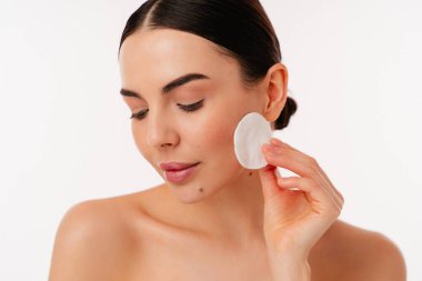 Yumuşak yüz temizleme ve sağlıklı cilt bakımı için pamuk pedi kullanan dingin bir bireyi gösteriyor. Minimalist bir geçmişe sahip temizlik, güzellik ve kişisel bakımı vurgular.