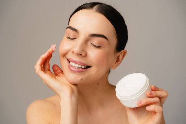 Gülümseyen bir şahsın konteynırını tutarken cilt bakımı yaptığı yakın plan. Güzellik, özgüven ve kişisel sağlık kavramlarını sakin ve tatmin edici bir ifadeyle gösteriyor..