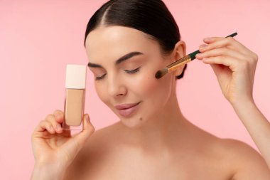 Sıvı temeli makyaj fırçası kullanarak uygulayan bir kişinin portresi, temiz, kozmetik odaklı estetik için pembe arka planlı doğal ve ışıl ışıl bir görünümü vurguluyor.