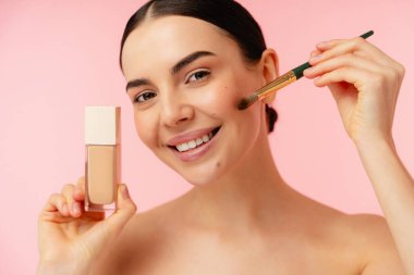 Elinde bir şişe sıvı fondöten ve makyaj fırçası ile gülümseyen bir birey, canlı pembe bir arka planla yanağına kozmetik uyguluyor, güzellik ve cilt kavramlarını vurguluyor..