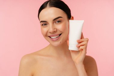 Stüdyo arka planında boş bir kozmetik ürün tüpü sergilerken gülümseyen ışıl ışıl bir insan..