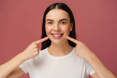 Beyaz gömlek giyen neşeli bir şahsın portresi, renkli bir arka planda gülümsemelerine işaret ediyor, pozitifliği, diş temizliğini, kendine güveni ve mutlu ve canlı bir görünümü temsil ediyor..