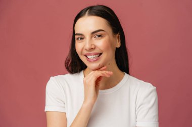 Pembe bir zemine poz veren neşeli parlak kız mutluluk ve pozitiflik yayıyor. Temiz beyaz bir tişört giyerek, sıcaklığı ve yakınlığı yansıtıyorlar, bu görüntüyü yaşam tarzı için ideal hale getiriyorlar..