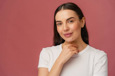 Basit beyaz gömlekli gülümseyen bir şahsın portresi, pembe bir zemine karşı poz veriyor, elini hafifçe kaldırmış, sıcaklığı, güveni ve yaklaşımı yayıyor..