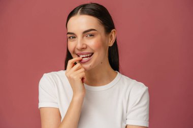 Beyaz tişört giyen, neşeli bir ifade sergileyen, neşeli bir jest yapan, sade pembe bir arka plana dayanan bir kişinin portresi..