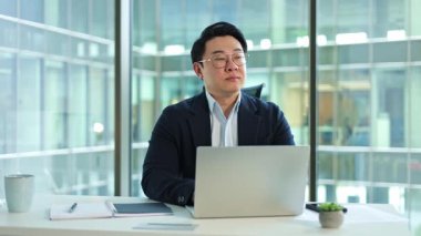Odaklanmış Koreli işadamı geniş pencereli modern bir ofiste dizüstü bilgisayarla çalışıyor. Yeni bir proje düşünüyor..