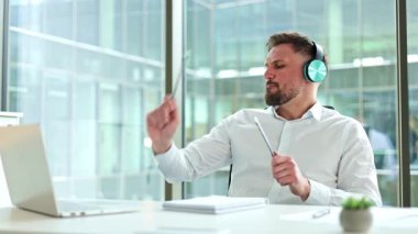 Neşeli iş adamı kulaklık takıyor, kalem ve not defterini ofis masasında davul olarak kullanıyor. Müzikten hoşlanan adam.