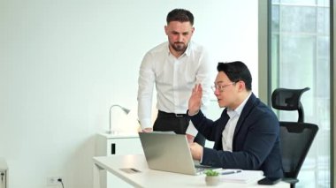 Modern ofiste birlikte çalışan ciddi iş adamları. Koreli iş adamı ve meslektaşı dizüstü bilgisayar kullanıyor ve belgeleri tartışıyorlar. İş toplantısı, takım çalışması, iletişim.