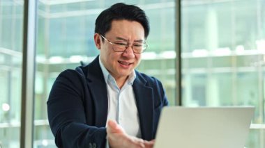 Asyalı sinirli iş adamının sorunları var. O, ofiste masada oturuyor, dizüstü bilgisayarını kullanıyor ve belgeleri okuyor. Stresli ve sinirli..